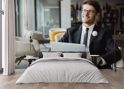 Portrait of happy young businessman sitting on armchair with laptop computer in hotel hall Wall mural