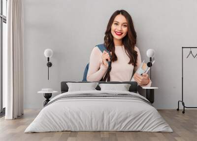 Portrait of intellectual female student with backpack being educated in university posing with book and laptop over gray wall Wall mural