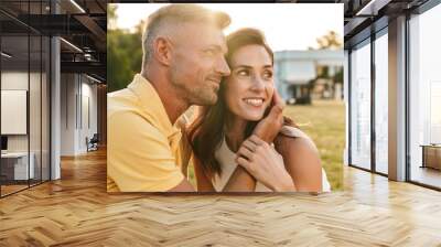 Portrait of lovely middle-aged couple looking aside at copyspace and hugging while sitting on grass in summer park Wall mural