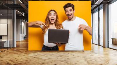 Portrait of pleased positive man and woman holding and pointing fingers at black laptop, isolated over yellow background Wall mural