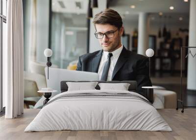 Portrait of pleased young businessman sitting on armchair with laptop computer in hotel hall Wall mural