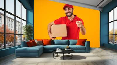 Portrait of positive delivery man in red uniform pointing finger at camera and holding packaging box Wall mural