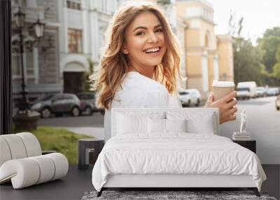 portrait of smiling european woman strolling through city street with silver laptop, and takeaway co Wall mural