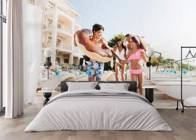 Portrait of smiling kids and parents walking near swimming pool, and carrying rubber ring outside hotel during vacation Wall mural