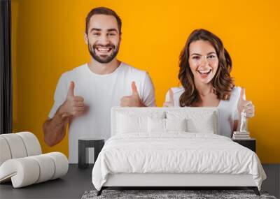 Portrait of smiling people man and woman in basic clothing showing thumbs up, while standing together isolated over yellow background Wall mural