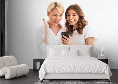 Portrait of two amazed surprised women mother and daughter holding and using black smartphone with surprise, standing isolated over white background Wall mural