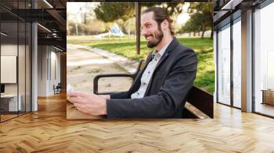 Profile portrait of happy businessman in suit smiling, while sitting on bench in city park and reading newspaper Wall mural