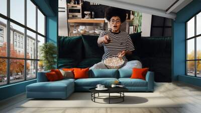 Shocked african woman watching tv and eating popcorn while sitting on couch Wall mural