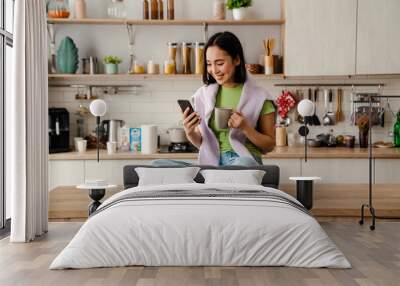 Smiling asian woman using mobile phone and drinking coffee while sitting in kitchen Wall mural