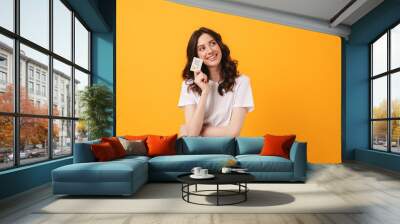 Smiling dreaming young woman posing isolated over yellow wall background holding debit card. Wall mural