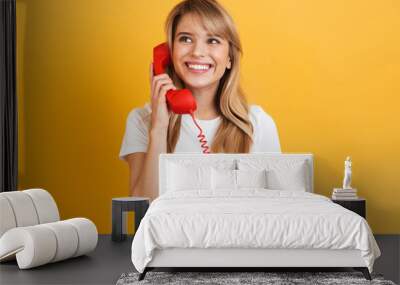 Smiling emotional happy young blonde woman posing isolated over yellow wall background dressed in white casual t-shirt talking by telephone. Wall mural