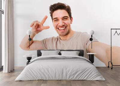 Smiling guy with brown hair showing peace sign on camera while photographing himself, isolated over white background Wall mural