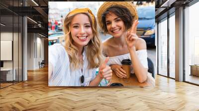 Smiling positive young women friends sitting outdoors in cafe drinking coffee take a selfie by camera. Wall mural