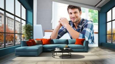 Smiling young man sitting and working with laptop at home Wall mural