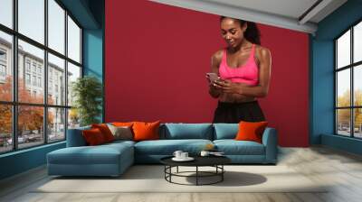 Strong young african fitness sports woman posing isolated over red wall background using mobile phone chatting. Wall mural