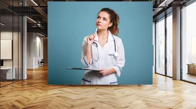 Thoughtful female doctor or nurse holding clipboard and looking up Wall mural