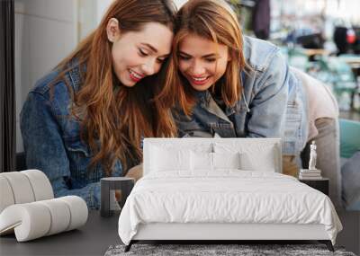 Two charming woman friends looking at mobile phone while sitting in cafeteria Wall mural