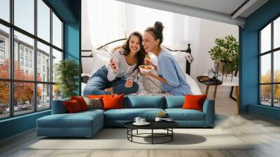 Two cheerful girls eating pasta and using mobile phone while sitting on bed together Wall mural