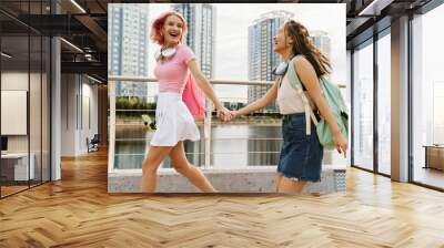 Two cheerful girls holding hands while walking together on city waterfront Wall mural