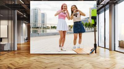 Two cheery girls recording videos with smartphone and showing heart gesture while standing on city waterfront Wall mural