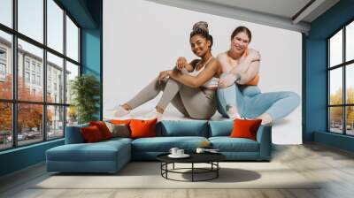 Two joyful women in sportswear posing together while sitting isolated over white wall Wall mural