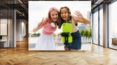 Two smiling girls recording videos with smartphone for their social media vlog on city waterfront Wall mural