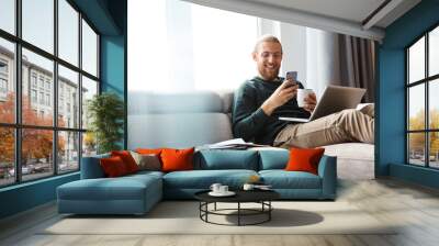 Young bearded man sitting in home using laptop computer and mobile phone. Wall mural