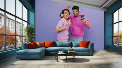 Young couple dancing while listening music with headphones and mobile phones isolated over purple background Wall mural