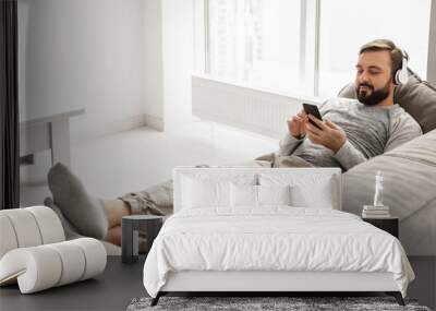 Young guy 30s in basic clothing lying on sofa at home, and listening to music on black mobile phone wearing headphones Wall mural