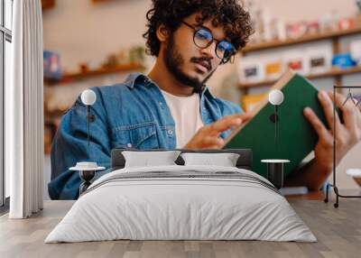 Young indian handsome man in glasses reading book at cafe Wall mural