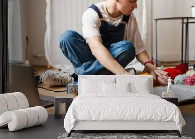 Young man using laptop and sewing machine while working on craft rug Wall mural