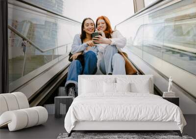 Young multinational women using cellphone while sitting on escalator Wall mural