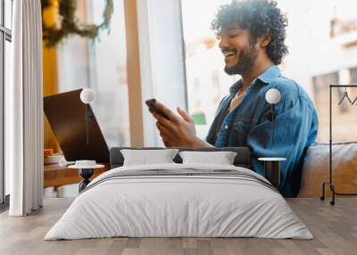 Young smiling handsome indian curly man looking phone while working Wall mural