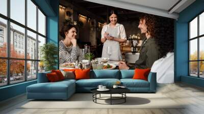 Young waitress serving group of cheerful friends in restaurant Wall mural