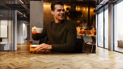 Young white man wearing sweater smiling while drinking coffee in cafe Wall mural