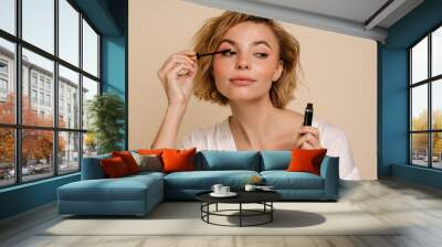 Young woman doing make up with mascara isolated over beige wall Wall mural