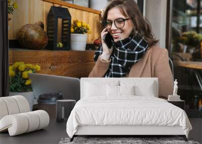 Young woman using laptop computer talking by mobile phone sitting in cafe outdoors. Wall mural