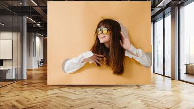 A sincere smiling young woman in sunglasses posing on a torn beige background. Wall mural