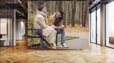 Friends, a senior and a young man sit in the park on a bench and talk in the autumn park. Wall mural