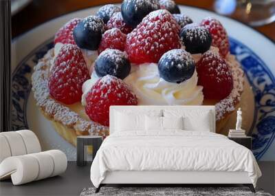 A close-up of a delicious pastry tart topped with fresh raspberries, blueberries, whipped cream and powdered sugar on a white plate with a blue and white rim. Wall mural