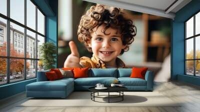 Cheerful child with curly hair giving thumbs up indoors, radiating positivity and joy with a brilliant smile Wall mural