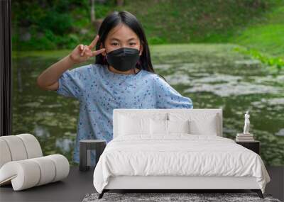 Portrait of Asian child girl posing cute act in a park left hand on waist. right-hand shows two fingers, black long hair, wearing a black color face mask Wall mural