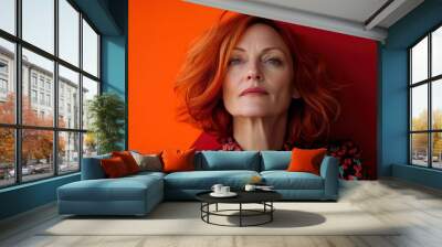 A mature woman with striking red hair and a thoughtful expression stands against a bold, two-tone orange and red backdrop, conveying strength and timelessness. Wall mural