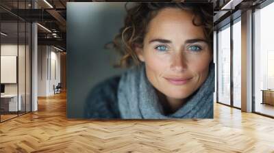 A woman with bright blue eyes and curly brown hair smiles warmly while wearing a cozy grey knitted scarf, capturing a serene and cheerful ambiance. Wall mural