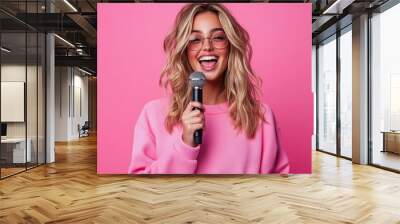 A young woman cheerfully sings into a microphone with a vibrant pink background, expressing joy and creativity through her stylish sunglasses and casual attire. Wall mural