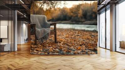 An inviting armchair is adorned with a warm blanket, basking in the golden hue of an autumn afternoon beside a gently flowing river, depicting perfect relaxation. Wall mural