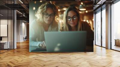 Two women wearing glasses huddle over a laptop in a brightly lit space, brainstorming ideas together, embodying teamwork and modern collaboration. Wall mural