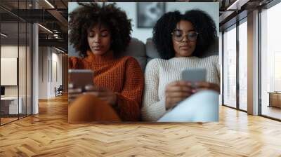 Two women with textured hair relax on a couch, immersed in their smartphones, showing the significance of digital connection and relaxation within a cozy home environment. Wall mural