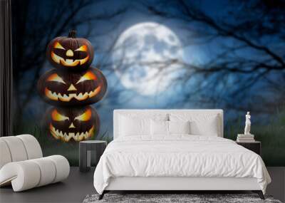 A pile of three spooky halloween pumpkin, Jack O Lantern, with an evil face and eyes on the grass with a misty night sky background with a full moon. Wall mural