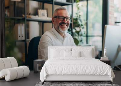 Portrait of a cheerful, bearded, and bespectacled older man with white hair, engaging with the camera with a bright smile while working at his computer in a sunny, modern office environment. Wall mural